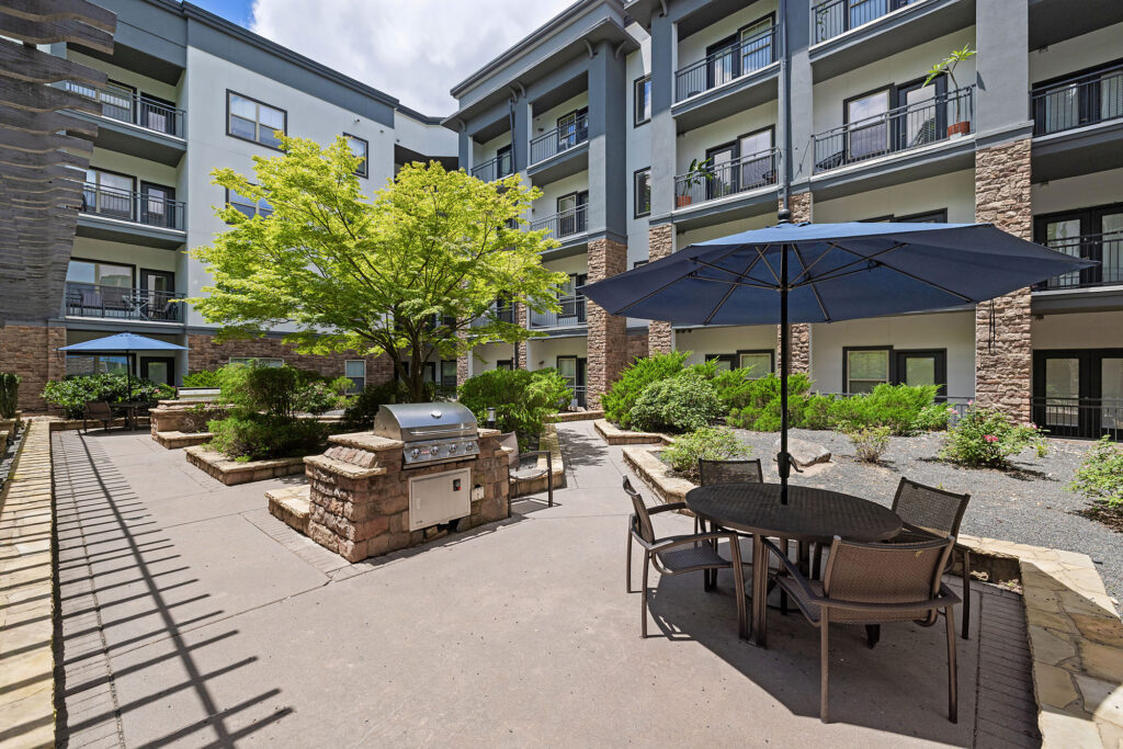 Outdoor patio area with a barbecue grill, round table and chairs under a large blue umbrella, surrounded by greenery and a modern apartment building with balconies in the background.