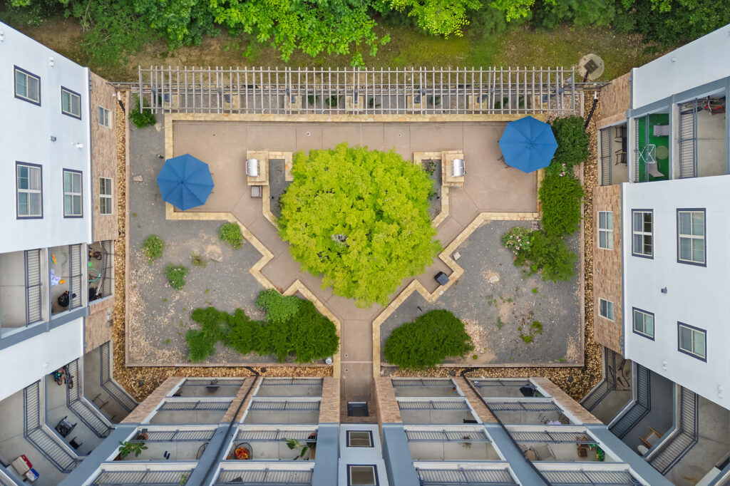 Aerial view of a building courtyard with a central green tree, symmetrical pathways, benches, two blue umbrellas, and surrounding apartment balconies. The courtyard is bordered by greenery and stone walls.