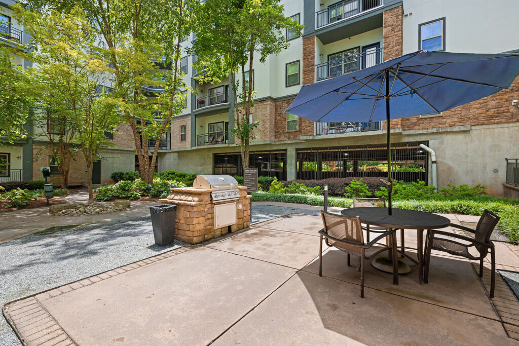Outdoor patio area with a round table, four chairs, and a large blue umbrella, next to a barbecue grill and trash bin. Surrounded by trees and greenery, with apartment buildings in the background.