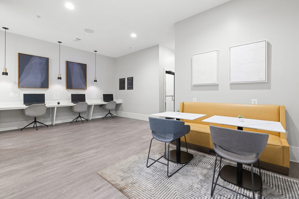 Modern workspace with a long built-in desk holding computers and chairs, pendant lights above, two abstract art pieces on the wall, and a lounge area with a yellow bench, gray chairs, and small tables.