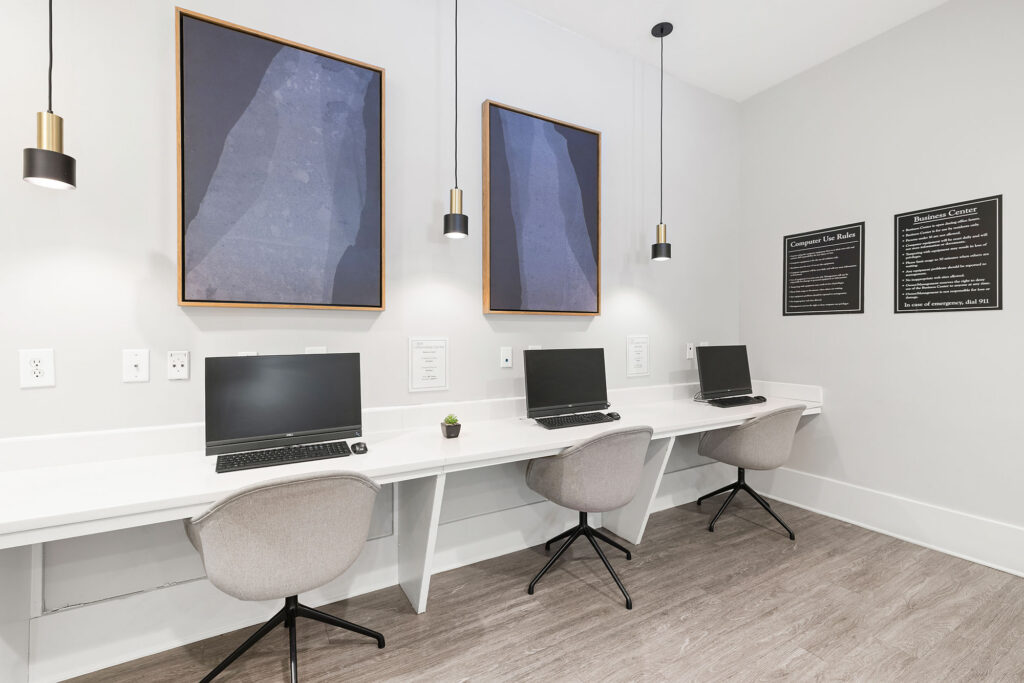 A modern computer lab with three desktop computers on a long white desk, gray swivel chairs, pendant lights, abstract wall art, and wood-style flooring.