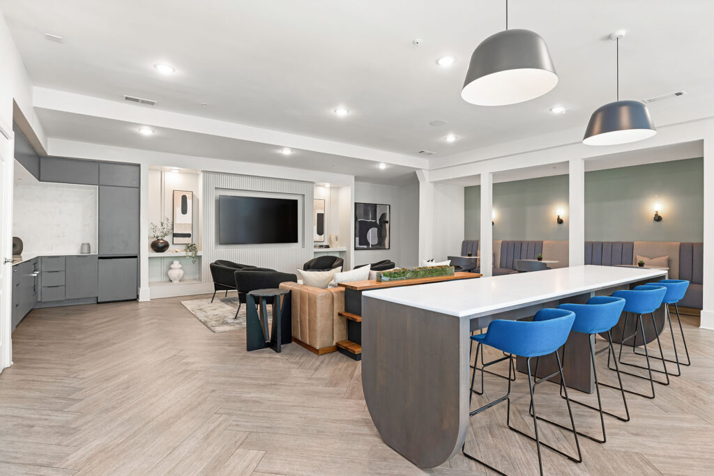 Modern communal lounge with a large TV, comfortable seating, a long table with blue stools, booth seating on the right, wood flooring, and stylish pendant lights in a bright, open space.