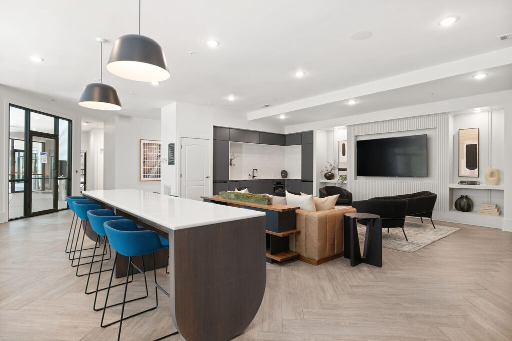Modern open-concept living area with a kitchen island and blue barstools, a cozy seating area with a TV, contemporary furniture, pendant lighting, and large windows letting in natural light.