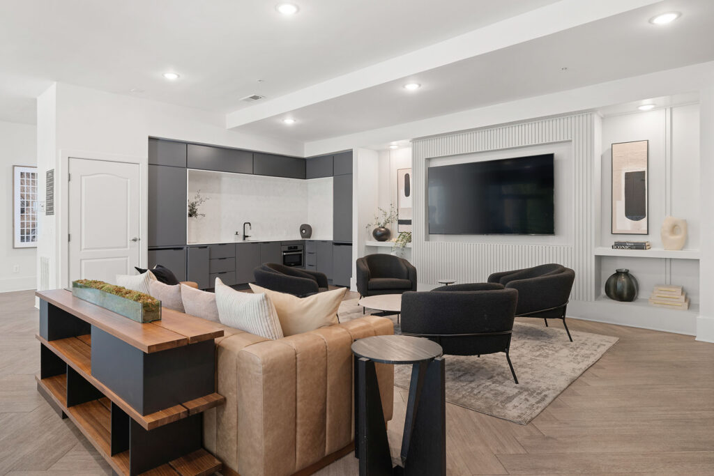 Modern living room with a tan leather sofa, black armchairs, built-in shelves, a large TV on the wall, a sleek kitchenette, wood flooring, and neutral decor in beige, black, and white tones.