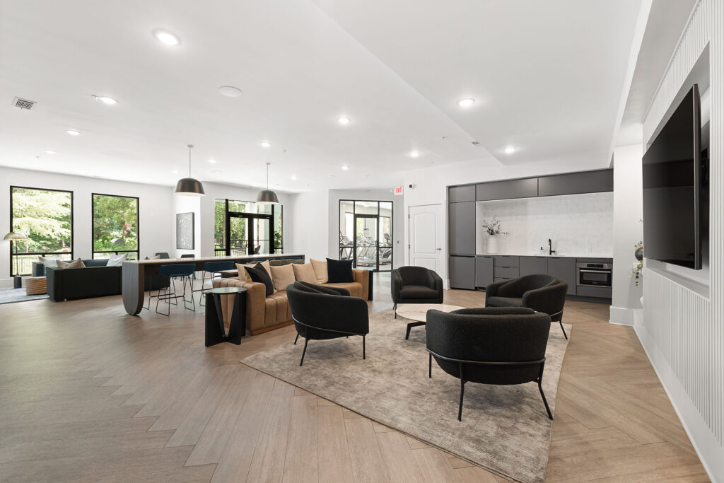 Spacious modern lounge with a seating area featuring a tan sofa, three black chairs, a coffee table, dining table, open kitchen, and large windows letting in natural light.