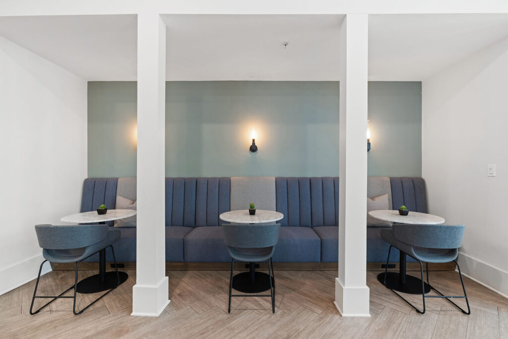 Modern seating area with two small round tables, blue upholstered chairs, and a long blue banquette against a light blue wall, illuminated by two wall sconces. The floor is light wood in a herringbone pattern.