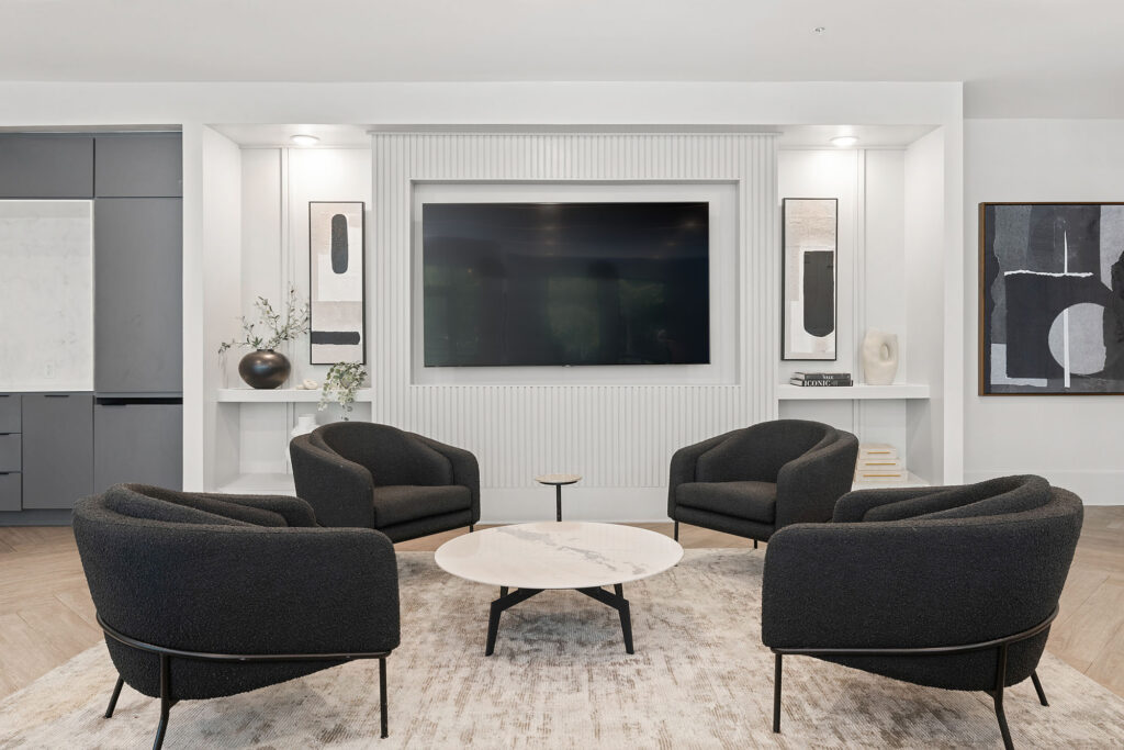 A modern living room with four black armchairs arranged in a circle around a round marble coffee table, a large wall-mounted TV, abstract artwork, and minimalist decor in neutral tones.