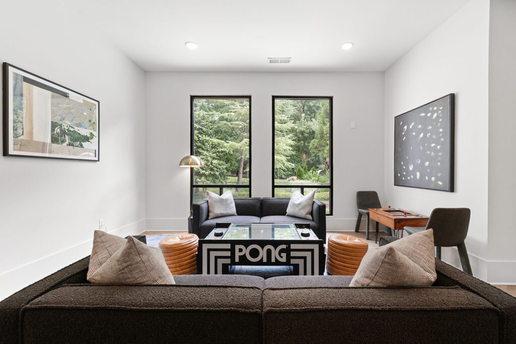 A modern living room with two dark sofas, a Pong-themed coffee table, two brown ottomans, large windows showing greenery, a desk with chairs, and contemporary wall art. The room has a clean, minimalist design.