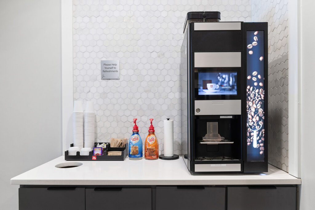 A modern coffee station with a coffee machine, disposable cups, wooden stirrers, sugar packets, creamers, and flavored syrups on a white countertop against a hexagonal tile backsplash.