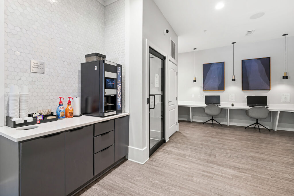Modern office break area with a coffee machine, cups, and syrups on a countertop. In the background, there is a glass door and a workspace with two computers, chairs, and abstract art on the wall.