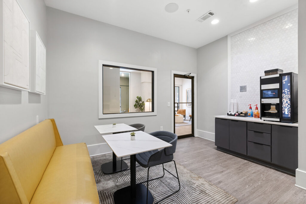 Modern break room with a yellow bench, two tables and chairs, a coffee machine on a counter, and a window into another room. The space features neutral colors and bright lighting.