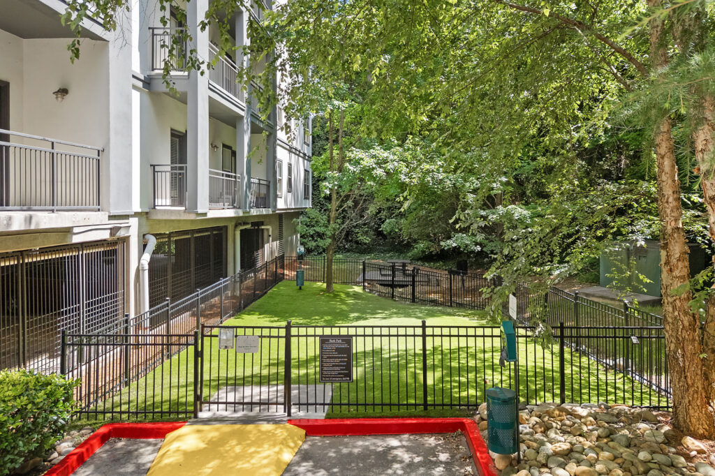 A fenced grassy area sits beside a modern apartment building, bordered by trees and a creek behind. The space features a pet waste station, a red-bordered area in the foreground, and a gravel walkway.
