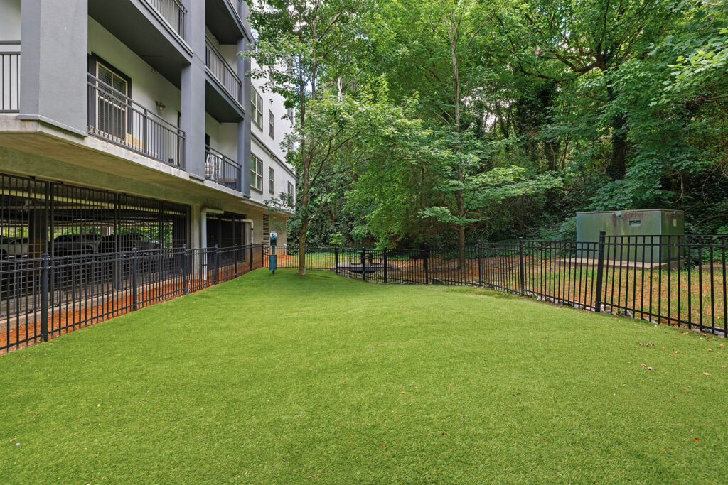 Fenced grassy area next to an apartment building with balconies, surrounded by trees and a shaded area with a bench. A utility box is visible in the background near the fence.