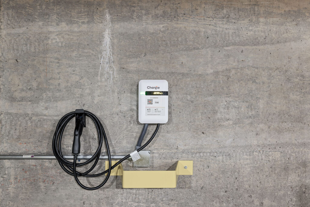 A wall-mounted electric vehicle charging station with a black charging cable coiled beside it, installed on a gray concrete wall with a yellow metal support underneath.