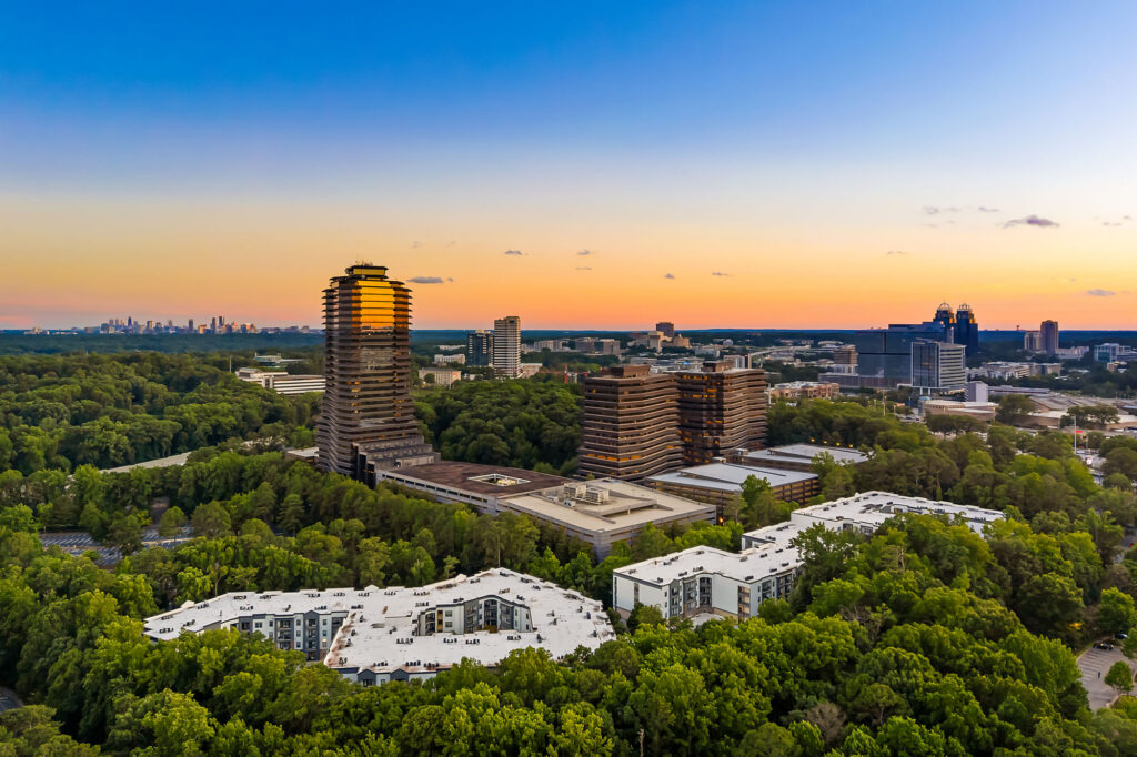 Aerial view of modern office buildings and apartments surrounded by dense green trees at sunset, with a city skyline visible in the distant background.