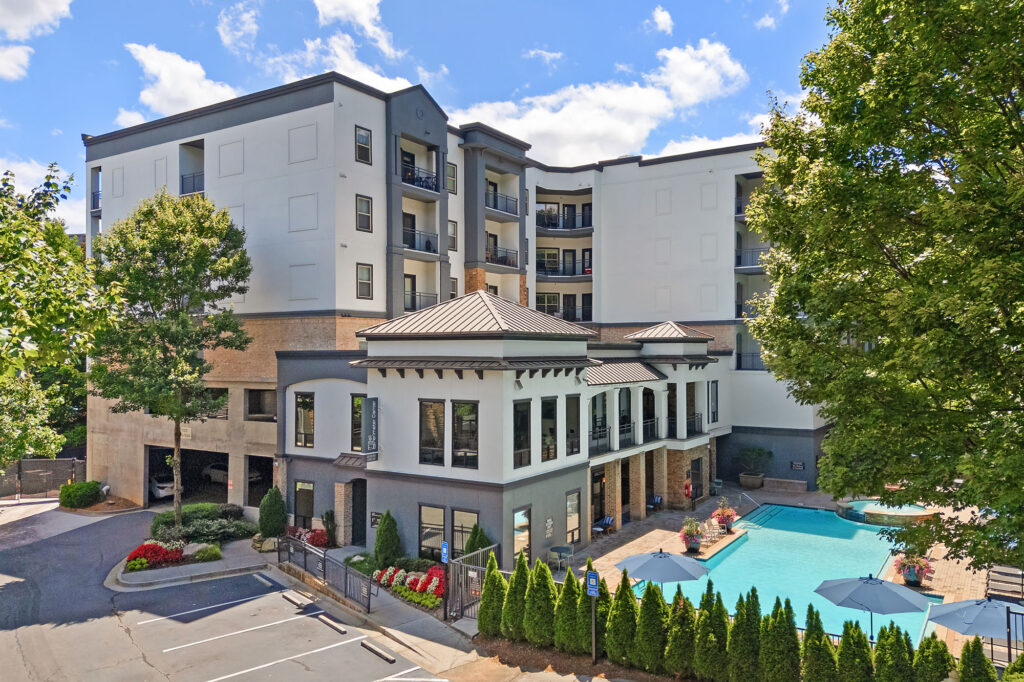 A modern, multi-story apartment building with balconies overlooks a swimming pool surrounded by lounge chairs and umbrellas. Trees and landscaped bushes decorate the area under a bright blue sky with scattered clouds.
