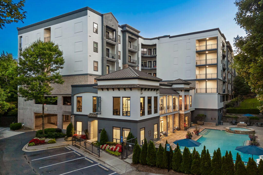 Modern apartment complex with large windows, balconies, and outdoor pool area surrounded by lounge chairs and umbrellas. The building is four stories tall and landscaped with trees and shrubs, seen at dusk with lights on.