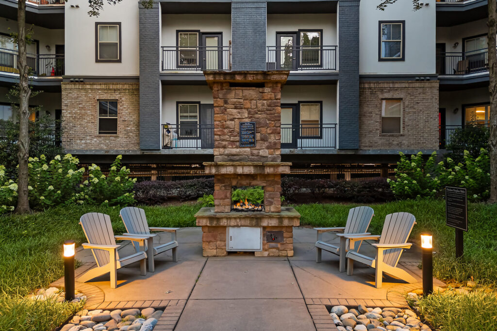 Four Adirondack chairs surround a stone outdoor fireplace on a paved patio, with apartment balconies and greenery in the background, illuminated by pathway lights at dusk.