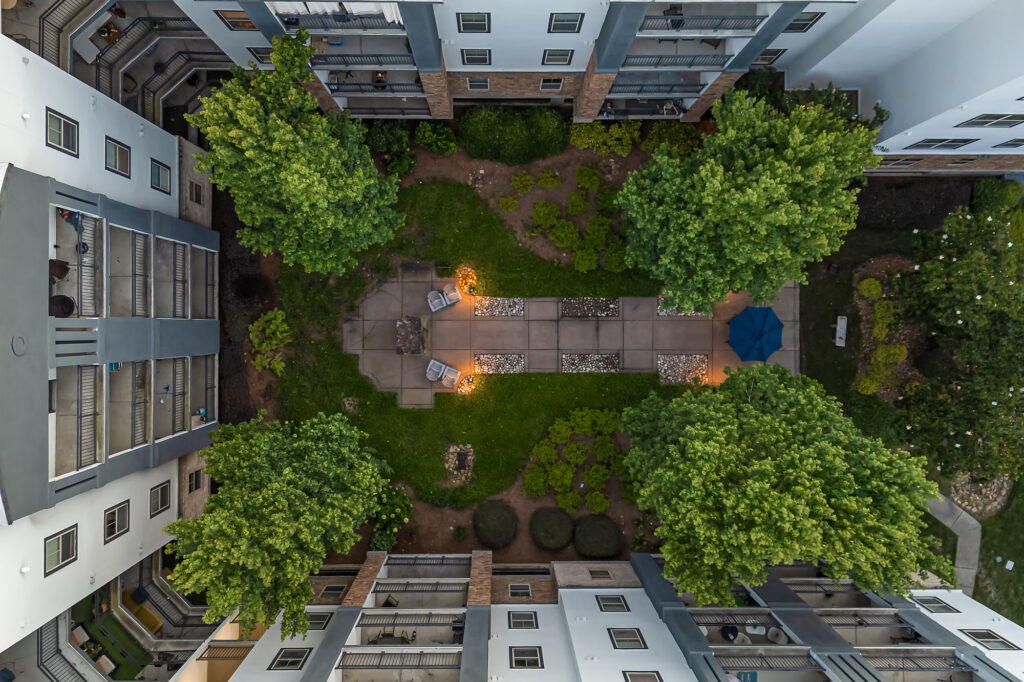 Aerial view of a courtyard between apartment buildings with green trees, walkways, patio furniture, and glowing outdoor lights at dusk.