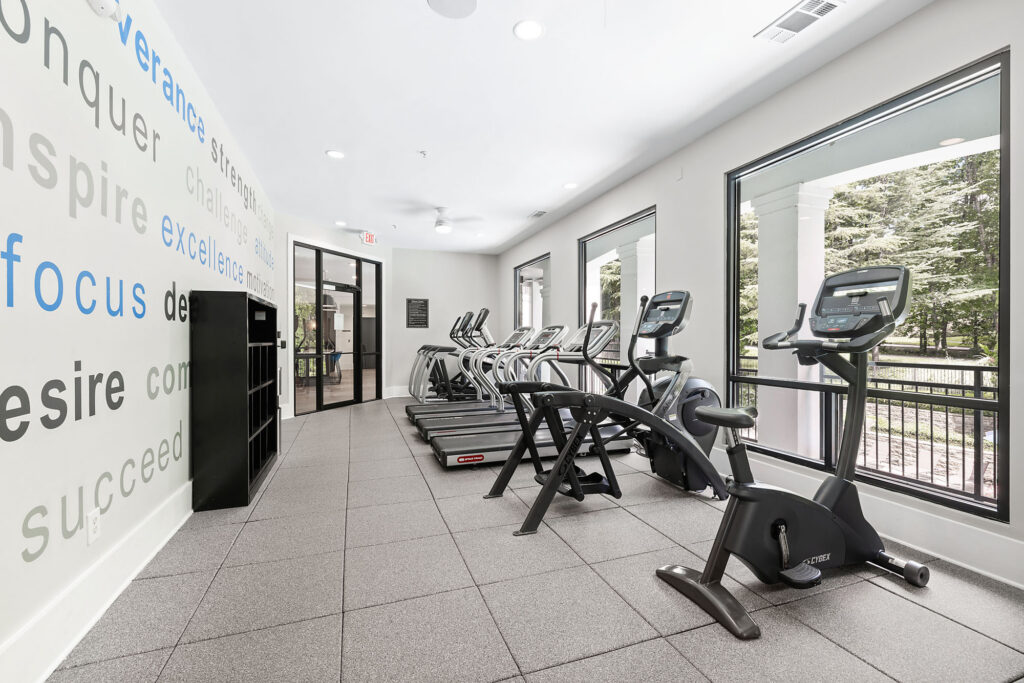 A modern gym with large windows, treadmills, and stationary bikes. The wall on the left has motivational words in blue and gray. Trees can be seen outside through the windows.