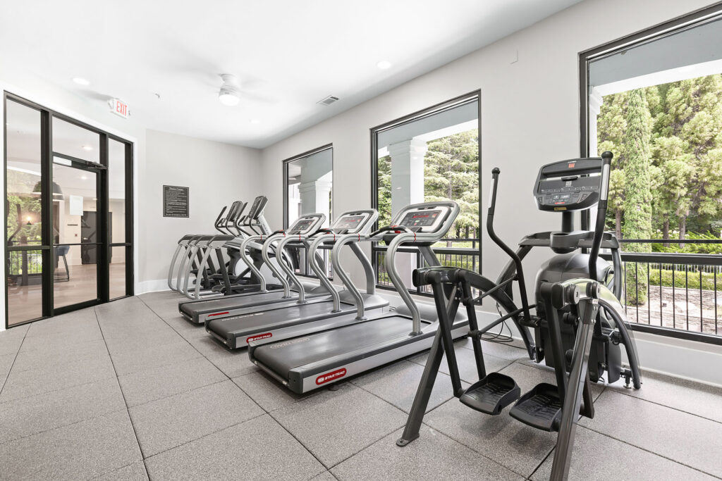 Modern gym with four treadmills and an elliptical machine facing large windows overlooking trees; bright, clean space with gray flooring and white walls.