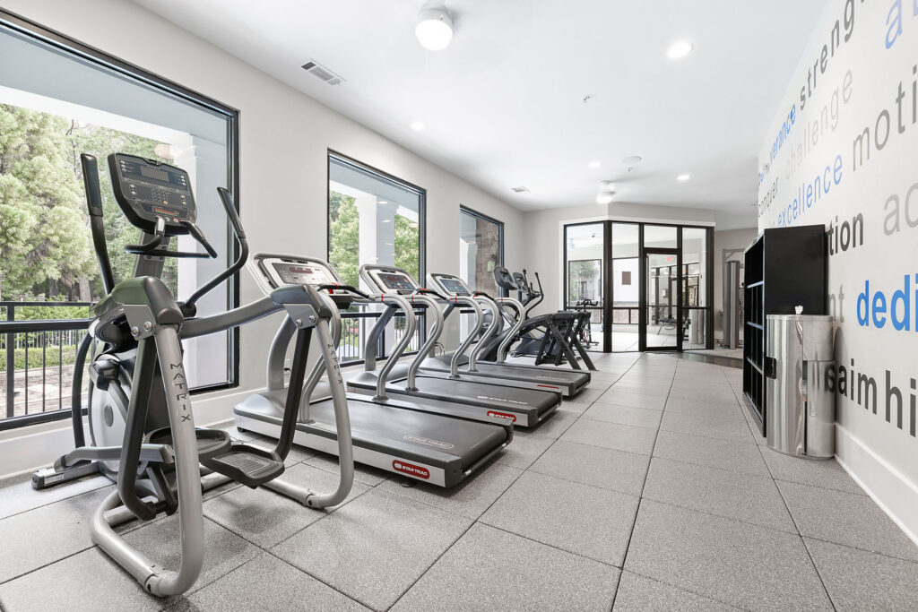 Modern gym with large windows, several treadmills, elliptical machines, and a shelf with towels. The walls are white with motivational words in blue and gray. Natural light fills the clean, spacious room.