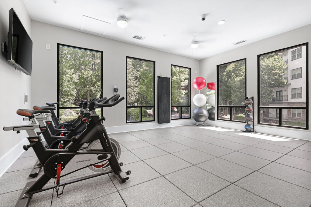 A bright fitness room with two stationary bikes, exercise balls stacked in a corner, free weights on a stand, large windows, and a wall-mounted TV. Trees and apartment buildings are visible outside.