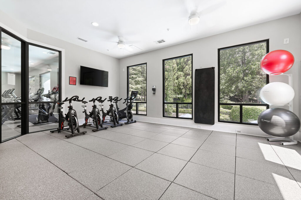 A bright, modern fitness room with three stationary bikes, a wall-mounted TV, exercise balls stacked in a corner, and large windows showing green trees outside.
