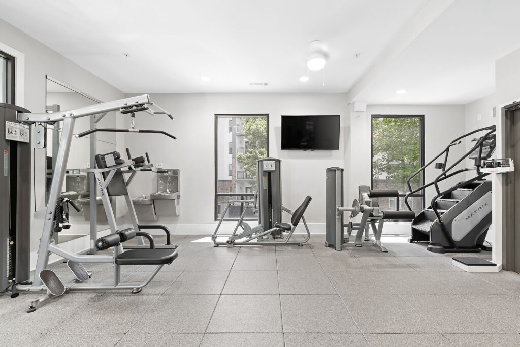 Modern gym with various exercise machines, including a weight station, cable machine, and stair climber. Large windows let in natural light, and a TV is mounted on the wall above the equipment. The space is clean and bright.