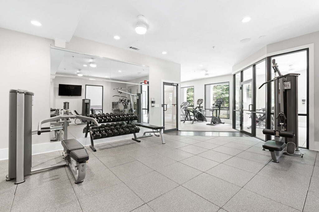 A modern gym with free weights on a rack, benches, strength training machines, and cardio equipment near large windows letting in natural light. The space has light walls and gray flooring.