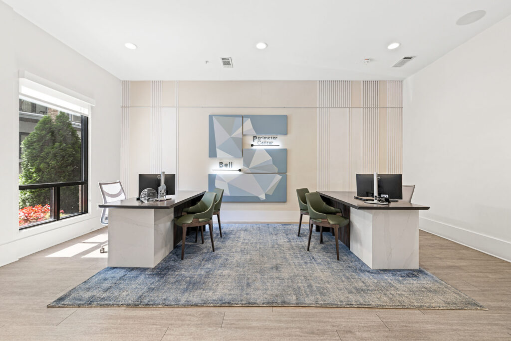 A modern office space with two desks facing each other, each with a computer and chairs. The room has a blue rug, large windows with natural light, and geometric wall art behind the desks.