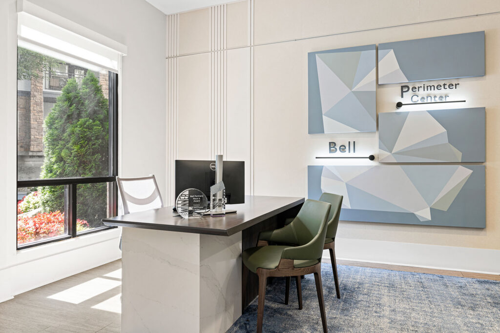 Modern office with a marble desk, two green chairs, a computer, and glass awards. Large window with view of trees. Abstract geometric wall art reads Bell Perimeter Center. Blue rug on wooden floor.
