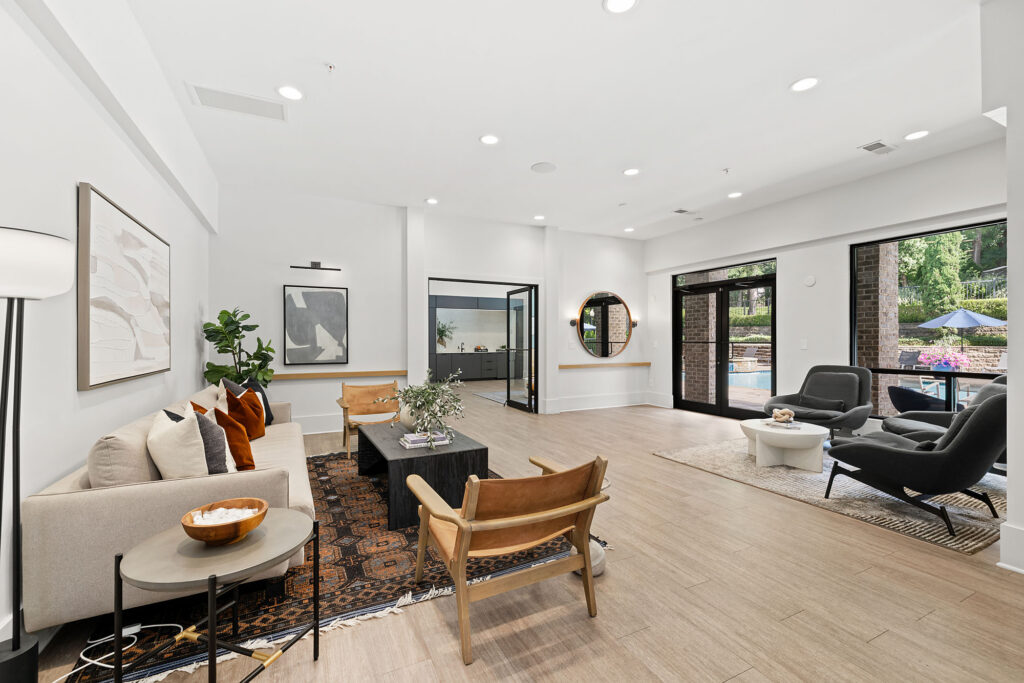 Modern, spacious living room with light wood floors, neutral furniture, large windows, and glass doors opening to a patio area. There are art pieces on the walls, plants, and a round mirror above a console table.