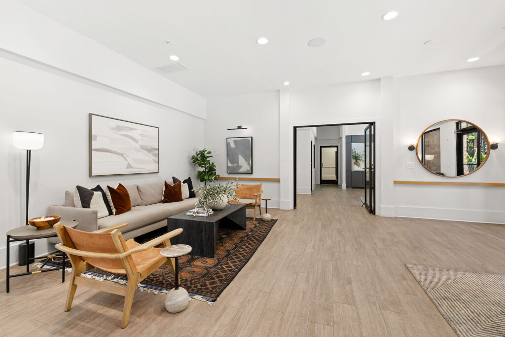 A modern, bright living room with a beige sofa, wooden chairs, a black coffee table on a patterned rug, abstract wall art, a floor lamp, and a round mirror. The space features light wood floors and white walls.