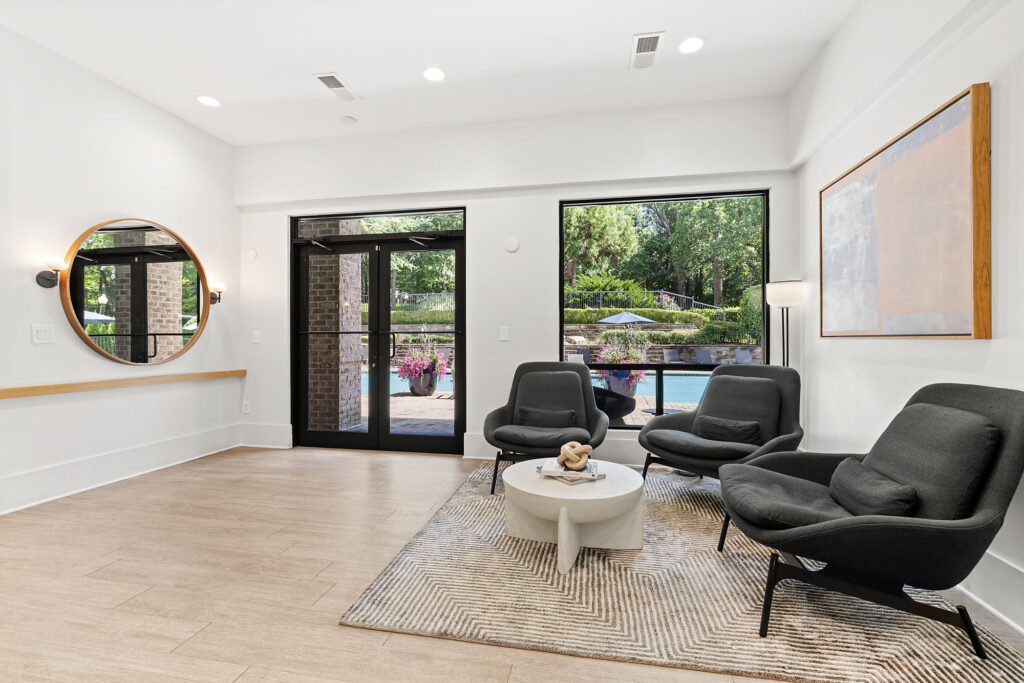 Modern lounge with three black chairs, a round white coffee table on a patterned rug, a large window and door overlooking a pool, a round wall mirror, and abstract wall art. The space is bright with natural light.
