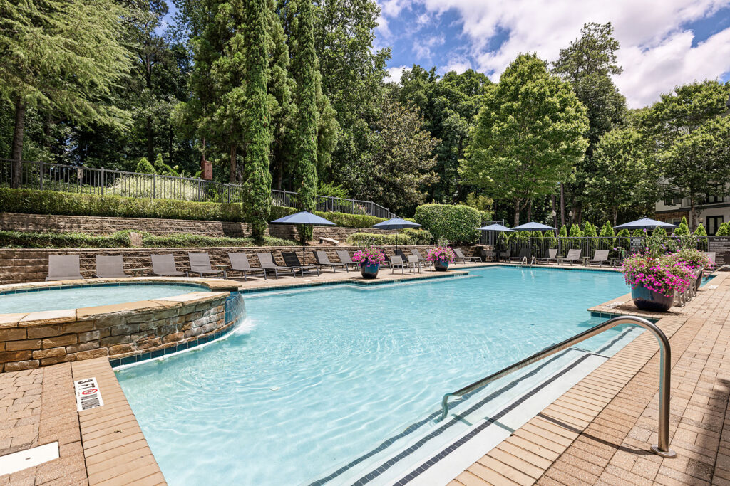 Outdoor swimming pool surrounded by lounge chairs and umbrellas, with a circular hot tub attached. The area is landscaped with trees, flowers, and greenery under a partly cloudy sky.