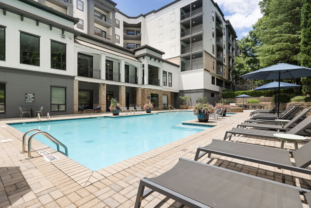 Modern apartment complex with a rectangular outdoor pool, surrounded by lounge chairs and umbrellas on a sunny day. Multi-story building and greenery are visible in the background.