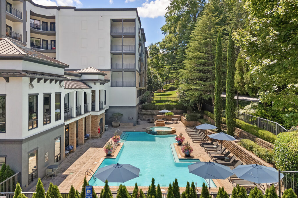 A modern apartment complex with a central outdoor swimming pool, lounge chairs, tables with umbrellas, landscaped greenery, and multi-story buildings in the background on a sunny day.