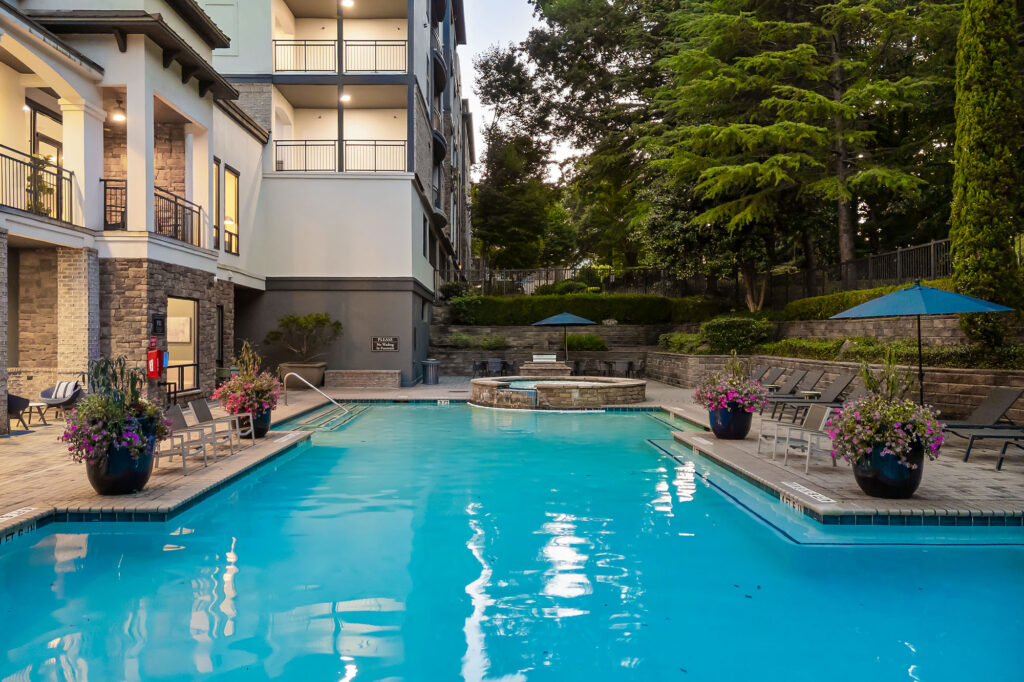 Modern apartment pool area at dusk, featuring clear blue water, lounge chairs, potted plants, and umbrellas. Trees and greenery surround the pool, and balconies overlook the space. A hot tub sits at one end of the pool.
