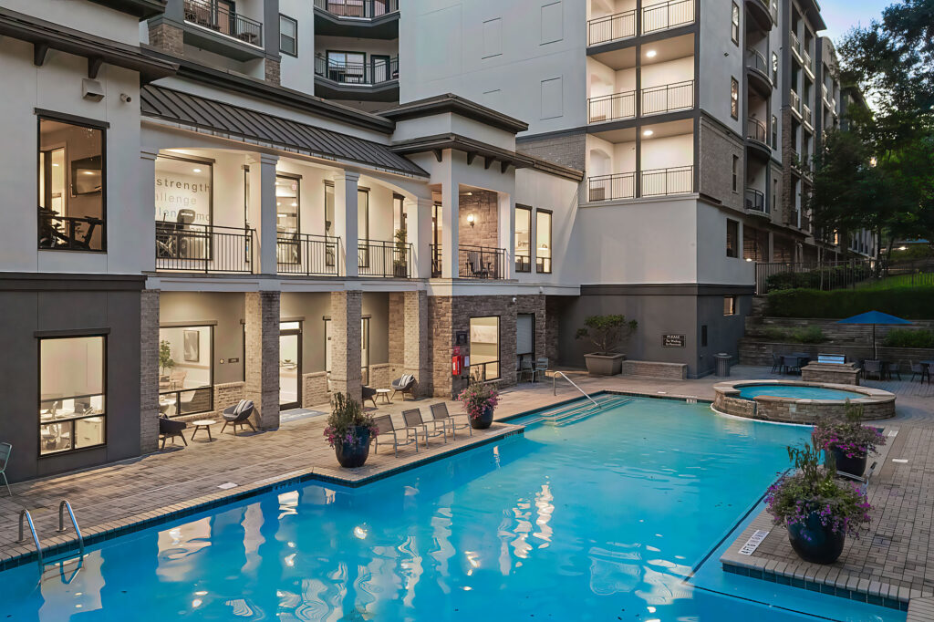 Modern apartment complex with large outdoor swimming pool, surrounded by lounge chairs and potted plants; adjacent hot tub; building with multiple balconies and large windows illuminated in the evening.