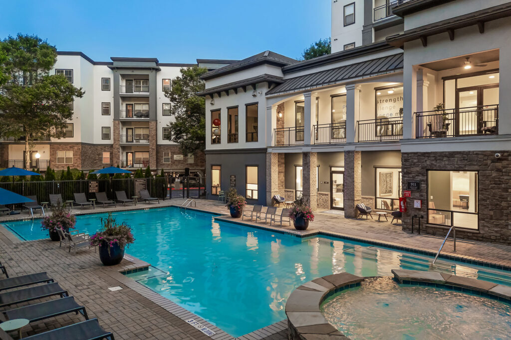 Modern apartment complex with a large outdoor pool, hot tub, lounge chairs, and balconies. The building has large windows, patio areas, and is surrounded by trees, with lights creating a welcoming atmosphere at dusk.
