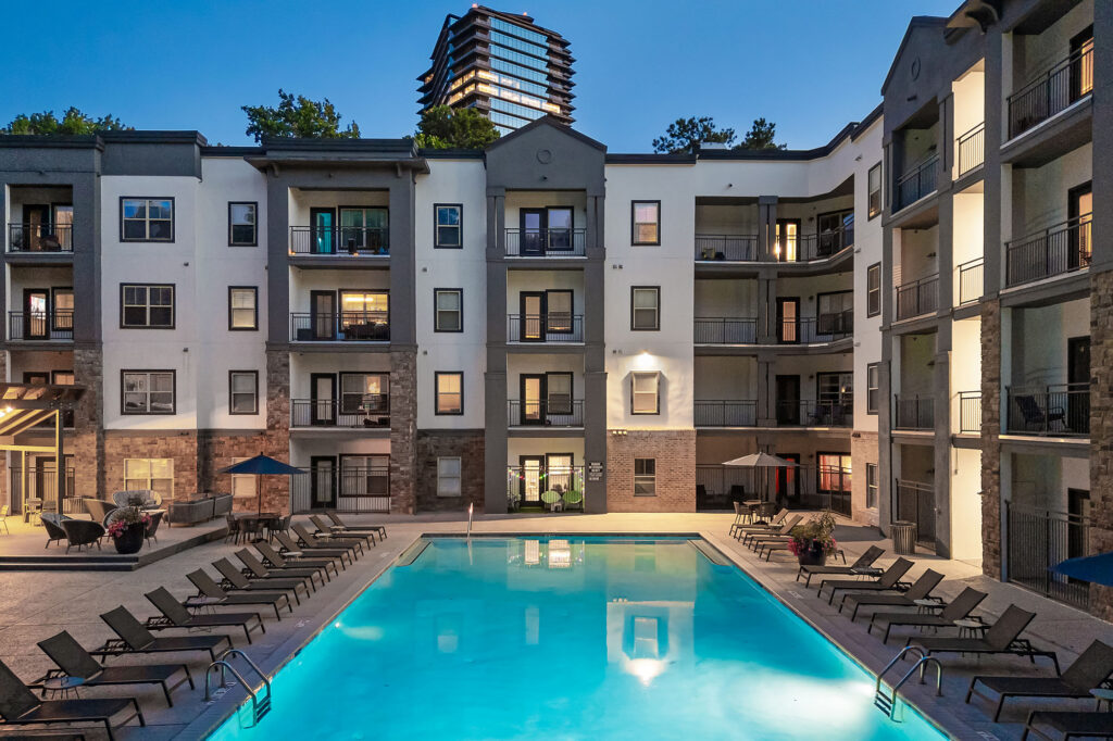 Apartment building at dusk with a lit outdoor swimming pool, lounge chairs, and balconies; a tall modern building is visible in the background surrounded by trees.
