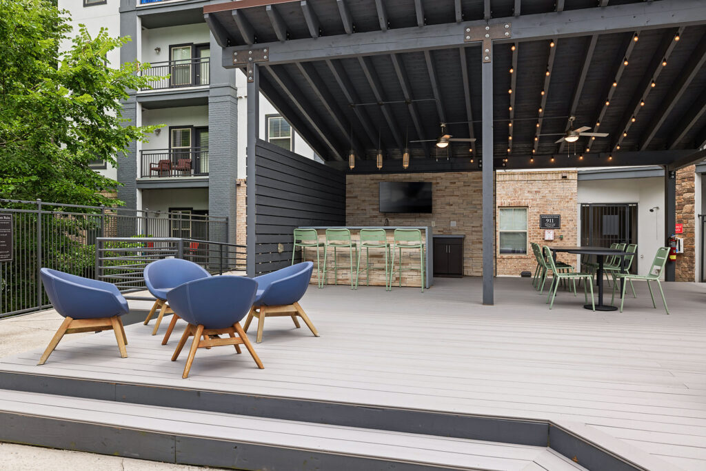 Outdoor covered patio with string lights, ceiling fans, a TV, high-top bar with stools, round dining table, and four modern blue lounge chairs. Modern apartment building and greenery are visible in the background.