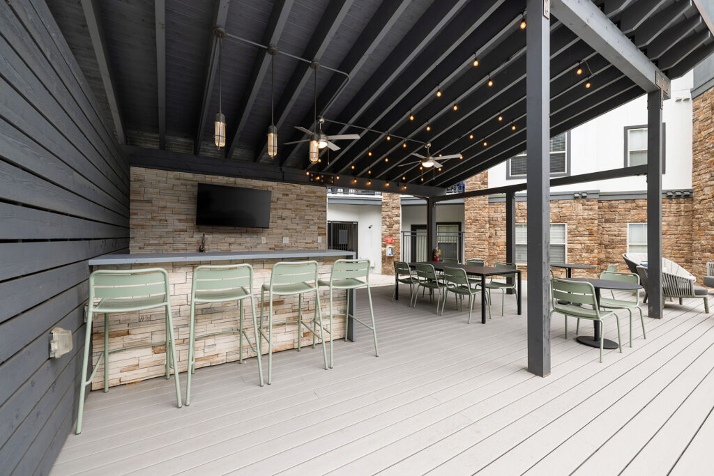 Covered outdoor patio with a bar counter and barstools, green chairs, dining tables, string lights, a wall-mounted TV, and a stone accent wall. The space has modern decor and light gray wooden flooring.
