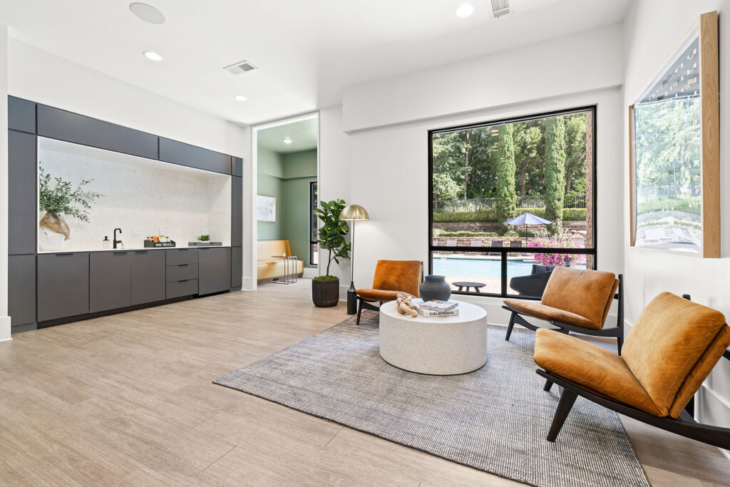 Modern lounge area with two brown chairs, a round white coffee table, and a gray rug near a window overlooking a pool and greenery. A kitchenette with dark cabinets and a potted plant are seen on the left.