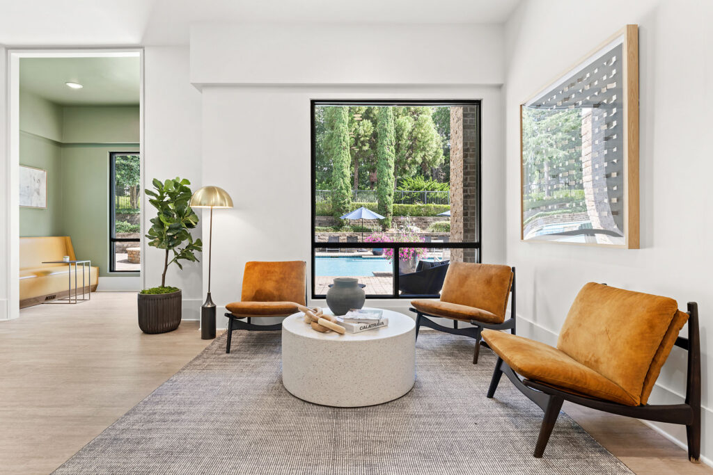 A modern lounge area with three tan chairs around a round white coffee table, a gray rug, a gold floor lamp, a potted plant, and a large window overlooking a pool and greenery. A framed artwork hangs on the wall.