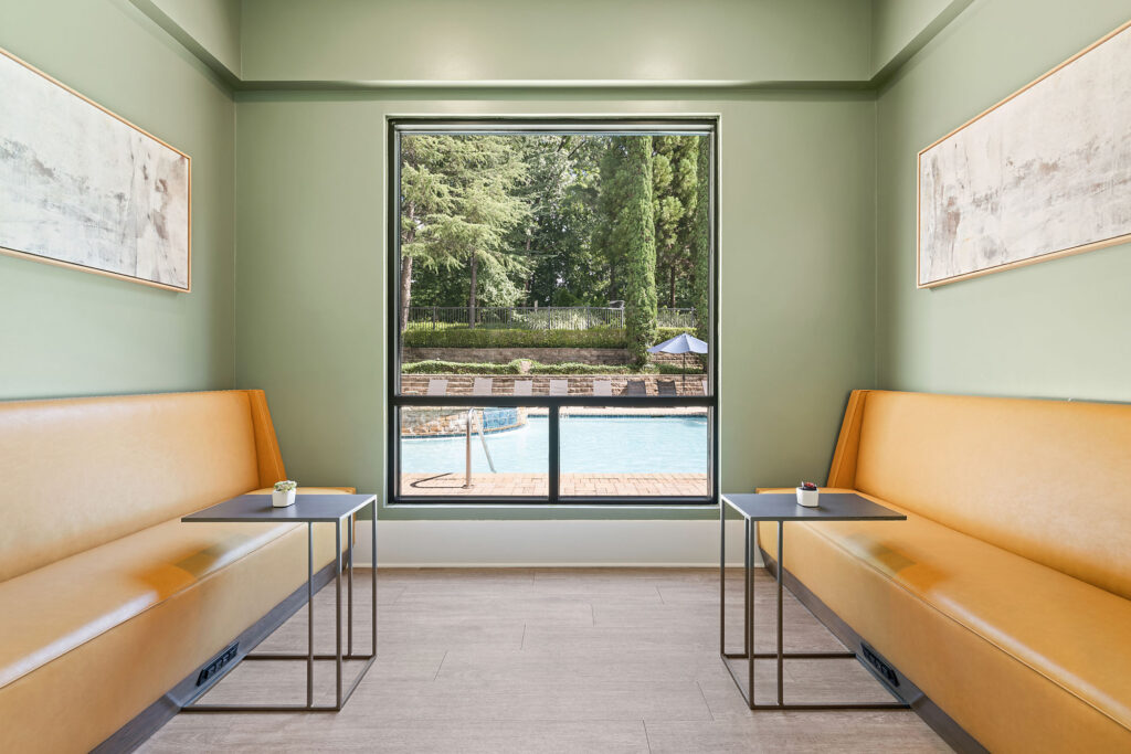 A modern seating area with two yellow benches and small tables faces a large window overlooking a swimming pool and trees outside. Abstract art hangs on the pale green walls.