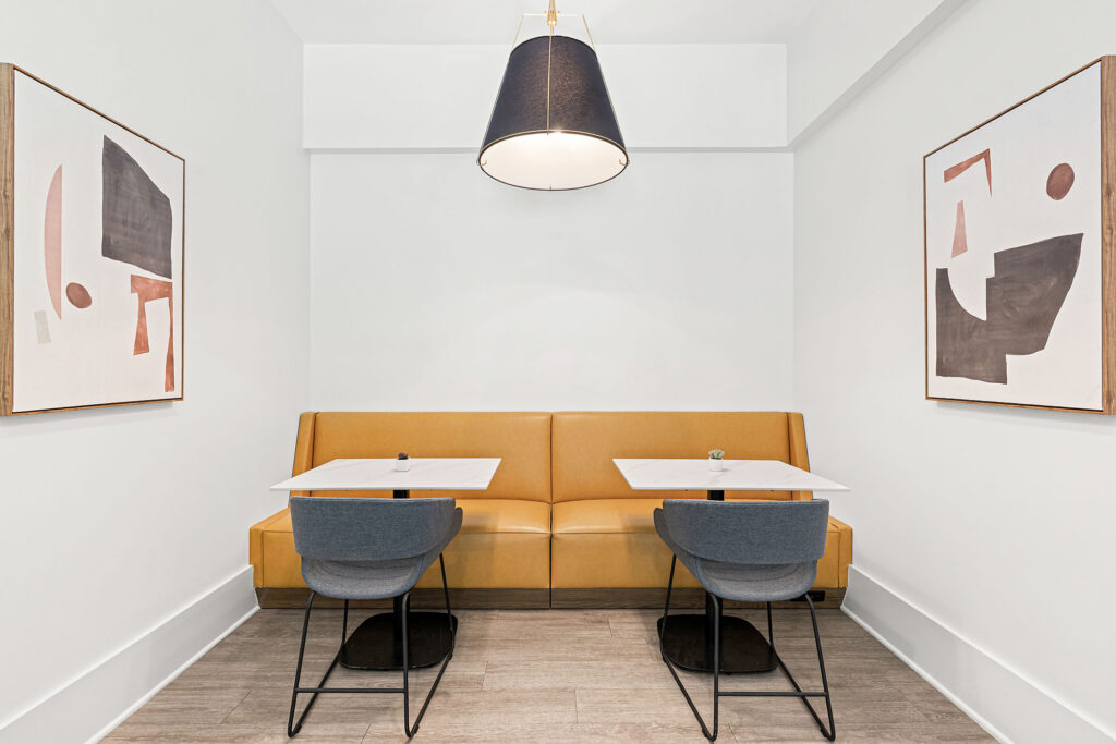 A modern, minimalist seating area with a mustard yellow banquette, two white tables, four gray chairs, abstract art on white walls, and a large black pendant light overhead.