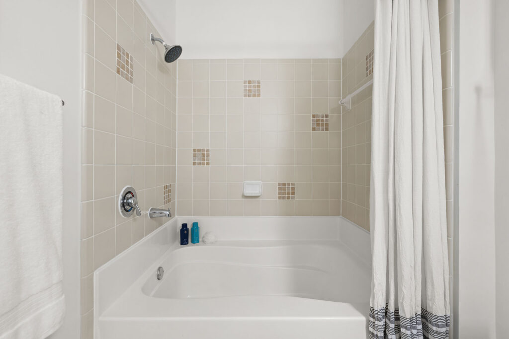 White bathtub and shower with beige tiles on the wall, some tiles patterned with small brown squares. A towel hangs on the left side, three bottles are on the bathtub ledge, and a striped shower curtain is on the right.
