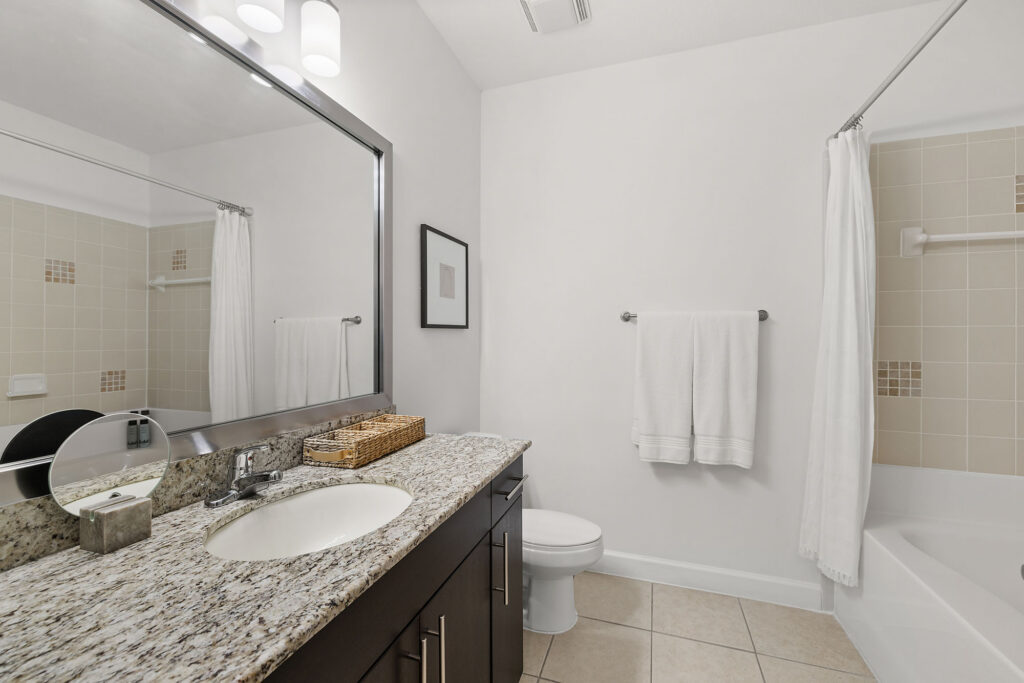 Modern bathroom with a granite countertop, sink, mirror, dark wood vanity, white towels, toilet, and a bathtub with a white shower curtain. Walls and tiles are light-colored, creating a clean and bright space.